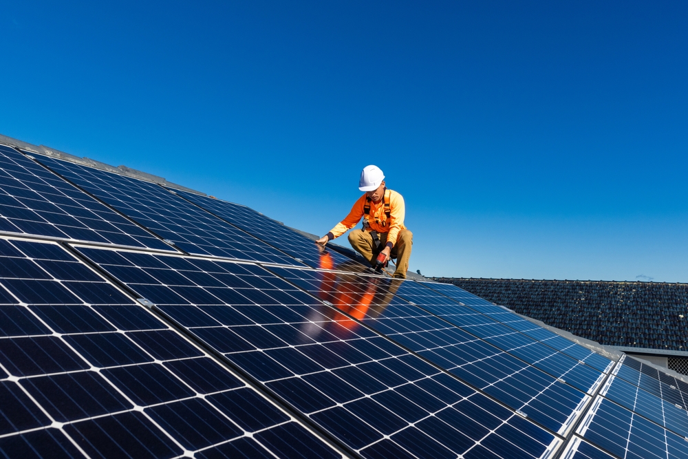 An engineer in a high-visibility vest and helmet inspects a vast array of solar panels under a clear blue sky.