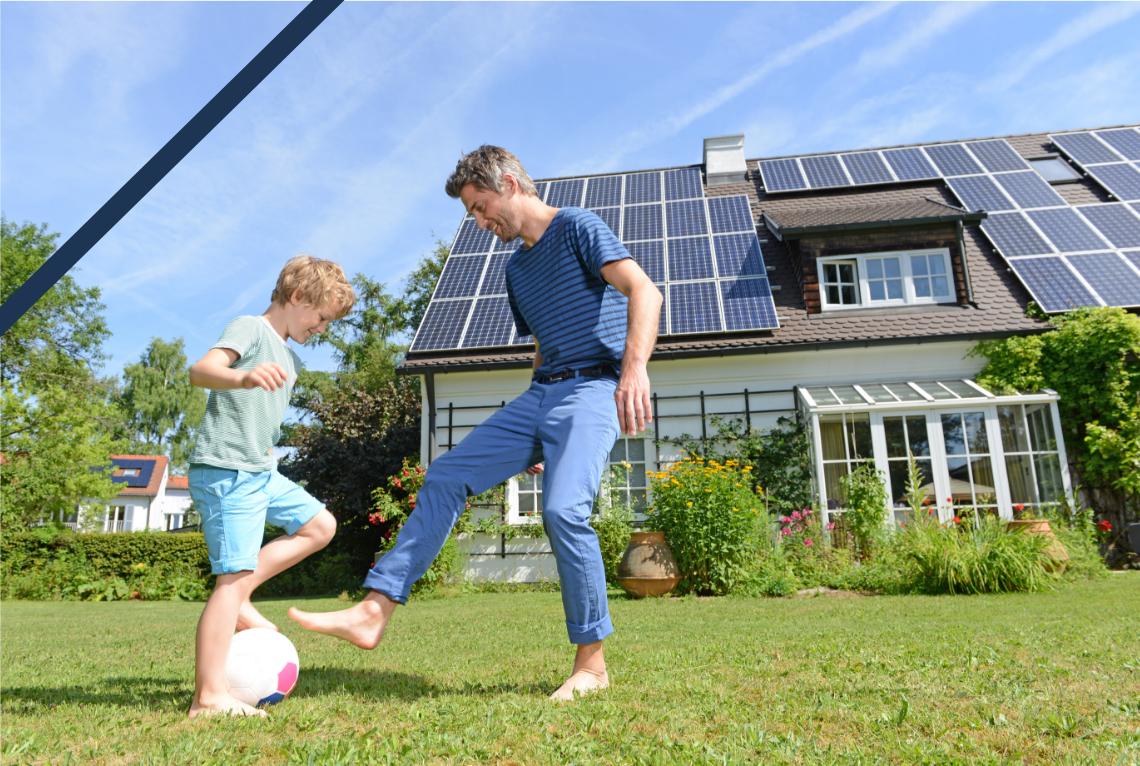 A man and a young boy playing soccer in a sunny backyard with a house equipped with solar panels in the background.