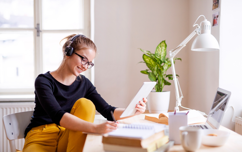 A joyful young woman wearing headphones while studying with a digital tablet at a bright home workspace.