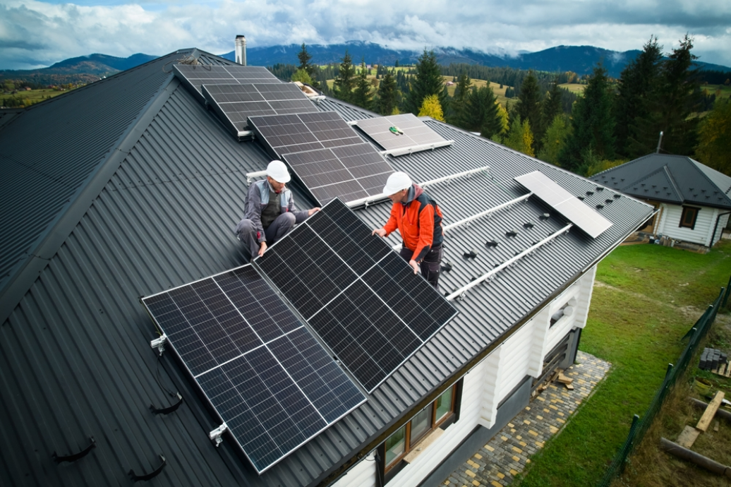 Workers installing a solar panel on the roof of a house with a scenic mountain landscape in the background.