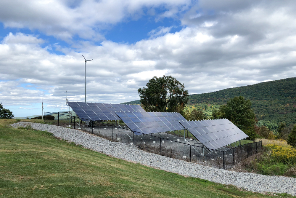 A series of solar panels sits on a gravel platform on a grassy hillside under a partly cloudy sky in New York. A small wind turbine is visible in the background, with trees and rolling hills extending towards the horizon.