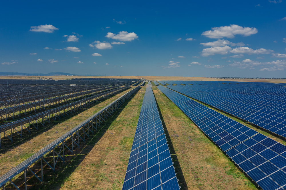 An expansive solar farm with numerous rows of blue photovoltaic panels stretching toward the horizon under a sunny sky with scattered clouds. The panels are mounted on metal frames, and the surrounding land is dry with some sparse vegetation, characteristic of modern solar farms.